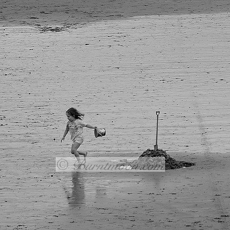 A black and white photo of a young girl running barefoot on a wet sandy beach, holding a small bucket. A sand mound with a spade stuck in it stands nearby, with reflections on the damp sand.