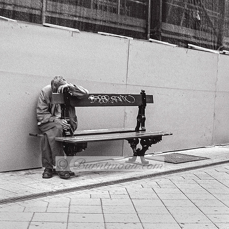 An elderly man sits slumped on a city bench in the rain, head resting on the bench's back, holding a bottle, surrounded by construction barriers in a gritty urban scene, captured in black and white.