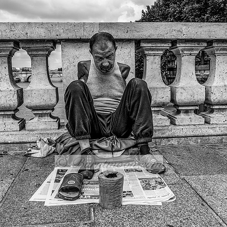 A physically disabled man sits on newspapers on a bridge over the Seine River in Paris, with a cup for donations in front of him, wearing mismatched sandals and a tank top, looking down in a solemn po