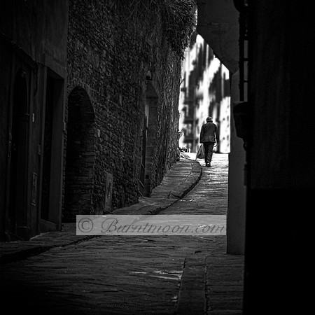 Black and white photograph of a solitary person walking up a narrow, cobblestone alley lined with ancient stone walls, with light streaming in from the background.