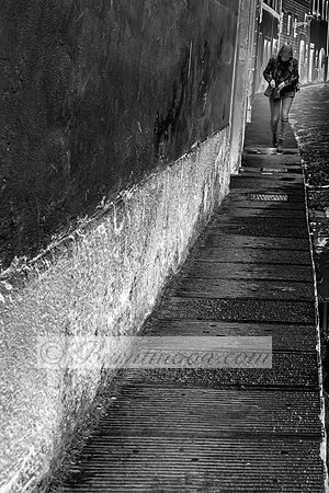 A black-and-white photograph of a hooded person walking along a narrow, worn sidewalk next to a rough-textured wall, evoking a sense of solitude and movement.