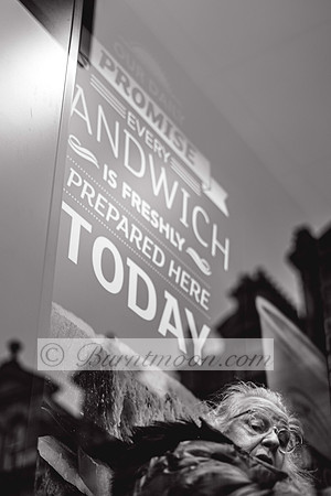 A low-angle, black and white photograph featuring a reflection of a large sign on a window in the upper left, which reads "OUR DAILY PROMISE EVERY SANDWICH IS FRESHLY PREPARED HERE TODAY". In the lowe