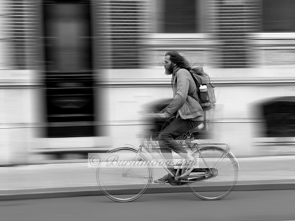 A black and white photograph of a person with a beard and backpack riding a bicycle on a street. The background, consisting of a building facade, is blurred horizontally to indicate motion, while the