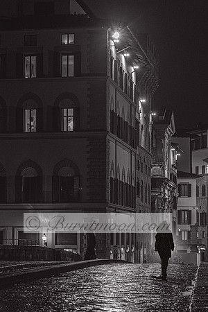 A black-and-white photograph of a solitary hooded figure walking along a wet cobblestone street at night, surrounded by historic buildings with glowing windows and dim streetlights.