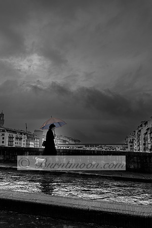 A black and white photograph of a person walking along a wet city street with a small dog, holding a colorful umbrella against a backdrop of stormy clouds and historic buildings.