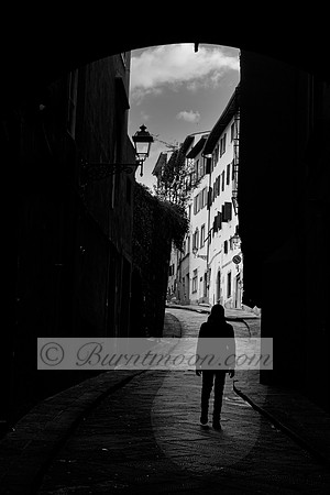 A silhouetted figure walks through a dark archway into a winding European street lined with old buildings, illuminated by soft daylight. The contrast between shadow and light enhances the mystery and