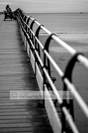 A black and white image of a person sitting on a bench on a pier, looking out at the ocean. The pier stretches into the distance, and the sea is calm.