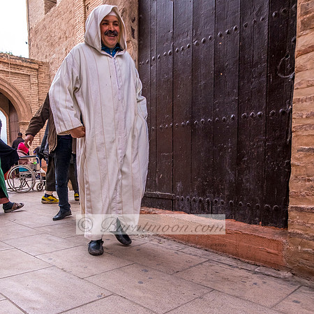 A man in a white djellaba walks past a large wooden door with a cheerful smile in Marrakesh, Morocco, near the Koutoubia Mosque, as people move through the historic street behind him.