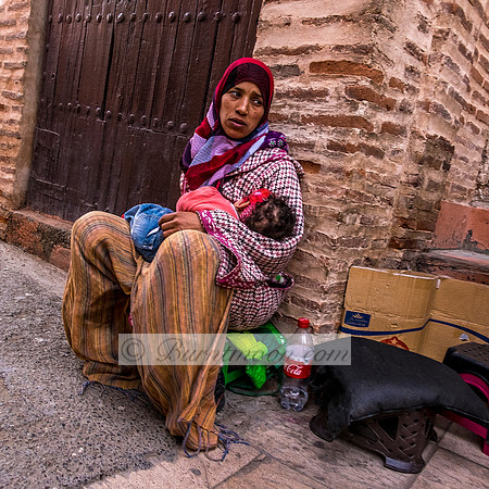 Woman in colorful clothing and hijab sitting on the ground, holding a sleeping baby, with brick walls and a wooden door in the background.
