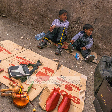 Two young boys in light purple jackets sit on cardboard boxes on a rough concrete ground, surrounded by scattered items including red bottles, a glass container, and shipping boxes, their expressions