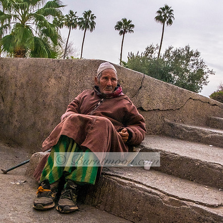 An elderly man wearing a brown djellaba and green-striped pants sits on worn stone steps in Marrakesh, Morocco, leaning against a cracked wall with a plastic cup beside him, while palm trees stand tal