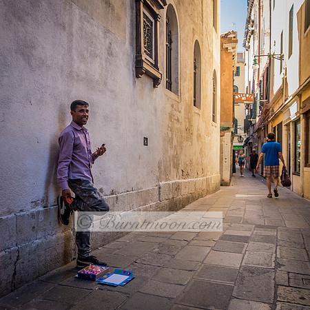 A man in a purple shirt stands against a stone wall in a narrow European street, with a small colorful display laid out on the ground in front of him, and pedestrians walking in the distance.