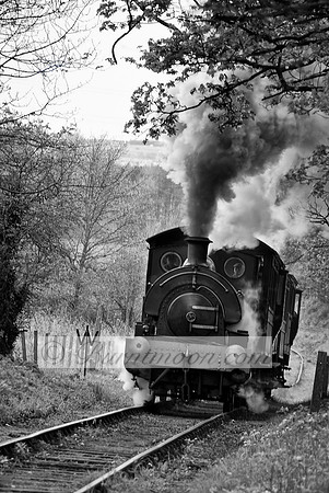 A black-and-white photograph of a vintage steam locomotive moving along a curved railway track, with thick smoke rising into the sky and trees framing the scene.