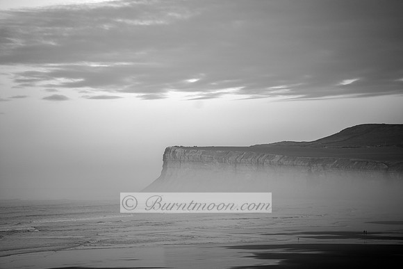 A black and white image of large cliffs shrouded in mist overlooking a turbulent sea. A small beach and a few tiny figures are visible at the bottom right.