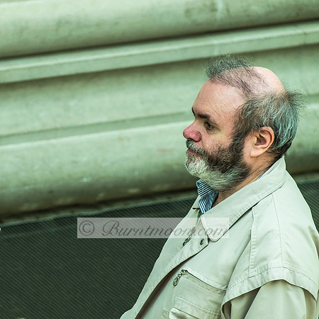 Side profile of a mature man with a gray beard, wearing a light cream jacket, standing against a sage green industrial background, looking thoughtfully to the side.