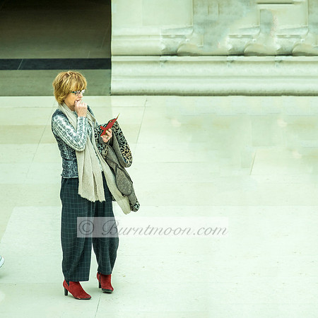 A middle-aged woman in a gray patterned sweater, checkered wide-leg pants, and red ankle boots stands alone in a light, spacious interior. She is looking down at something in her hand, with a contempl