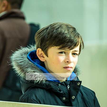 A young boy wearing a black winter coat with a blue hoodie and fur-lined hood, looking off to the side with a pensive expression against a blurred background.