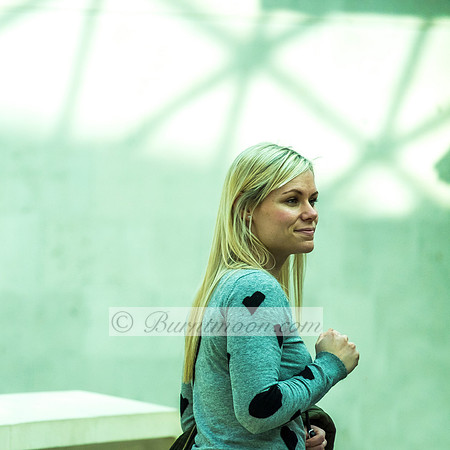 A blonde woman in a gray sweater with black heart patterns walks through a softly lit, modern space with a contemplative expression.