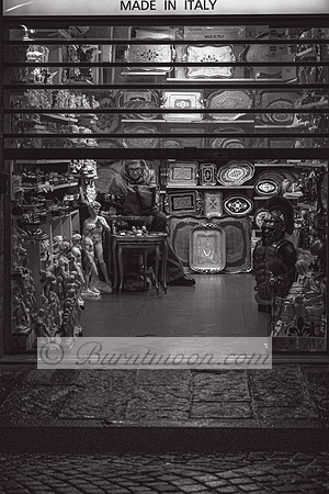A black and white photograph of an elderly shopkeeper sitting in a small, dimly lit antique store filled with ornate trays, classical sculptures, and various decorative objects, partially obscured by