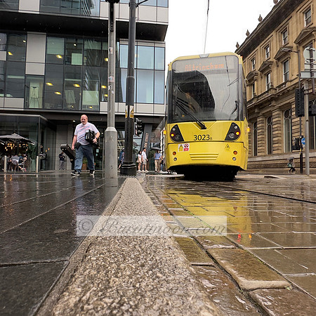 A yellow tram numbered 3023b on a wet city street, with modern glass buildings and classical stone architecture in the background, and a person standing near a street pole.
