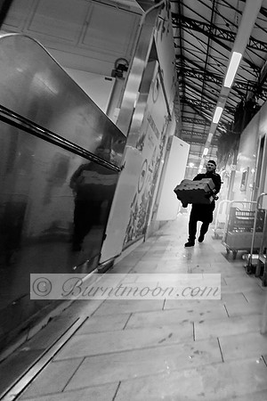 A black-and-white photograph of a worker carrying a heavy crate through a wet industrial corridor, with reflective surfaces and harsh overhead lighting.