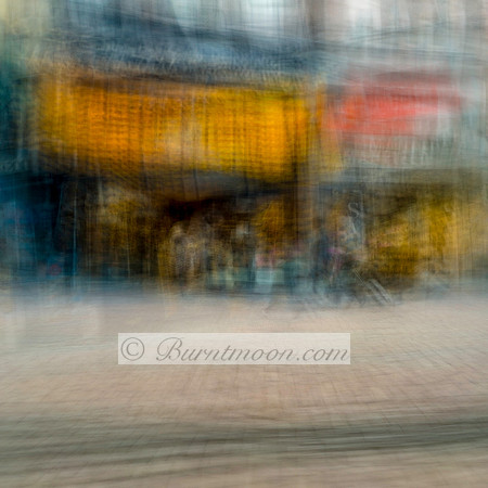 A blurred, abstract street scene in Amsterdam with yellow and orange storefronts and indistinct human figures created using intentional camera movement (ICM).