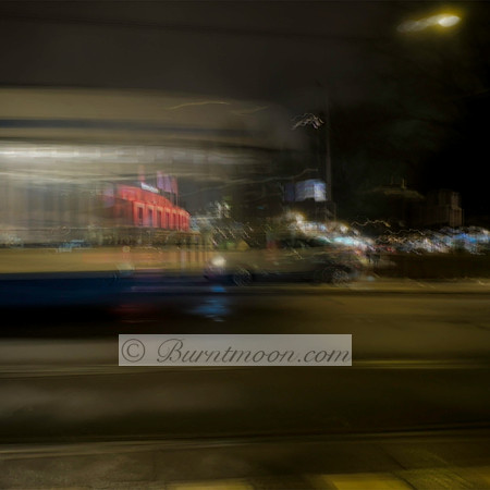 A blurred tram in motion passes through a city street at night in Amsterdam, with glowing lights, a red-lit building, and reflections on wet pavement creating an abstract, dreamy effect.