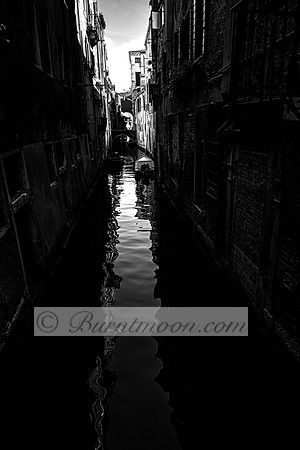 A narrow Venetian canal in black and white, with small boats moored along the edges and reflections of distorted light rippling across the water. A stone bridge in the distance frames the quiet, shado
