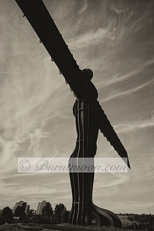 A black and white grainy silhouette of the Angel of the North sculpture against a textured cloudy sky.