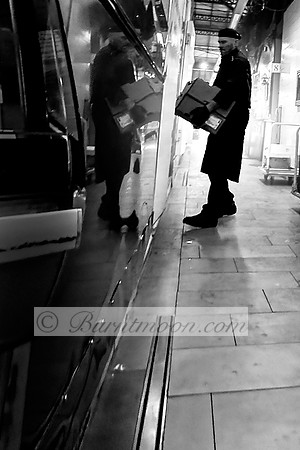 A black-and-white photograph of a worker carrying boxes in an industrial transit area, with his reflection visible on the side of a polished vehicle, emphasizing motion and labor.