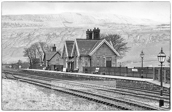 Black and white photograph of a quaint railway station in the Yorkshire Dales, with traditional architecture, lamp posts, and railway tracks leading into the distance.
