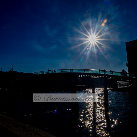 A photograph of silhouetted figures walking across a modern arched bridge with the sun shining brightly in a deep blue sky, casting shimmering reflections on the water below.