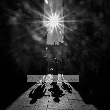A black-and-white photograph of three silhouetted people walking through a narrow alleyway, with the sun positioned directly above, casting long shadows on the ground and creating a radiant burst of l