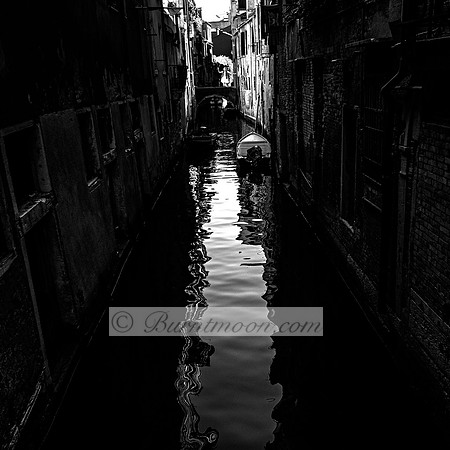 A narrow Venetian canal in black and white, with small boats moored along the edges and reflections of distorted light rippling across the water. A stone bridge in the distance frames the quiet, shado