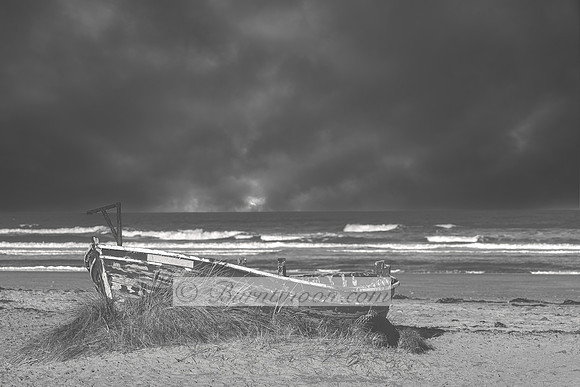 A black and white photograph of a decaying wooden boat on a sandy beach, surrounded by grass, with dark storm clouds and ocean waves in the background.