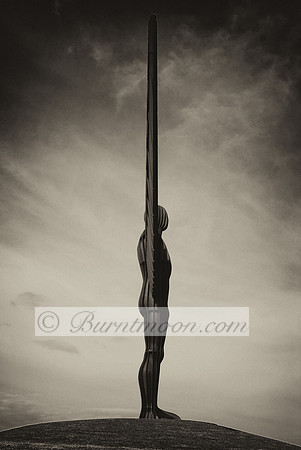 A black and white side-profile view of the Angel of the North sculpture, appearing as a thin silhouette against a textured cloudy sky.