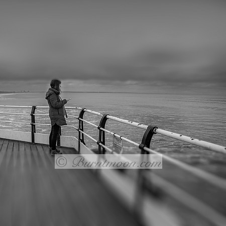 A woman in a winter coat stands on a weathered pier, gazing out at the ocean while holding her phone. The sky is overcast, and the foreground is blurred, emphasizing her solitude.