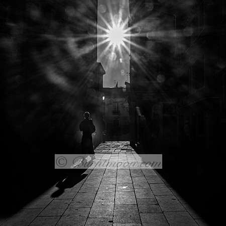 A black-and-white photograph of a lone silhouette walking down a narrow alleyway, with the sun creating a dramatic burst of light and long shadows on the stone pavement.