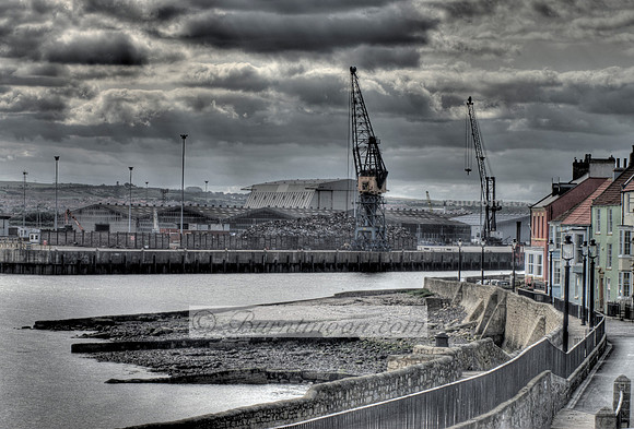 HDR photograph of Hartlepool Harbour with cranes, warehouses, and colorful houses along a waterfront path under a cloudy sky.
