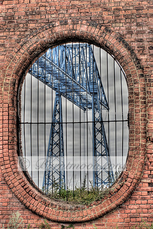 HDR photograph of the Transporter Bridge seen through a circular brick archway with iron bars, set against a cloudy sky.