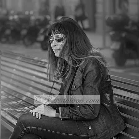 A woman in a denim jacket and sunglasses sits on a bench in an urban setting, gazing into the distance with a contemplative expression. The background is blurred, showing scooters and people in motion